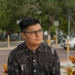A man with glasses sits in a park wearing a patterned shirt and using earbuds.